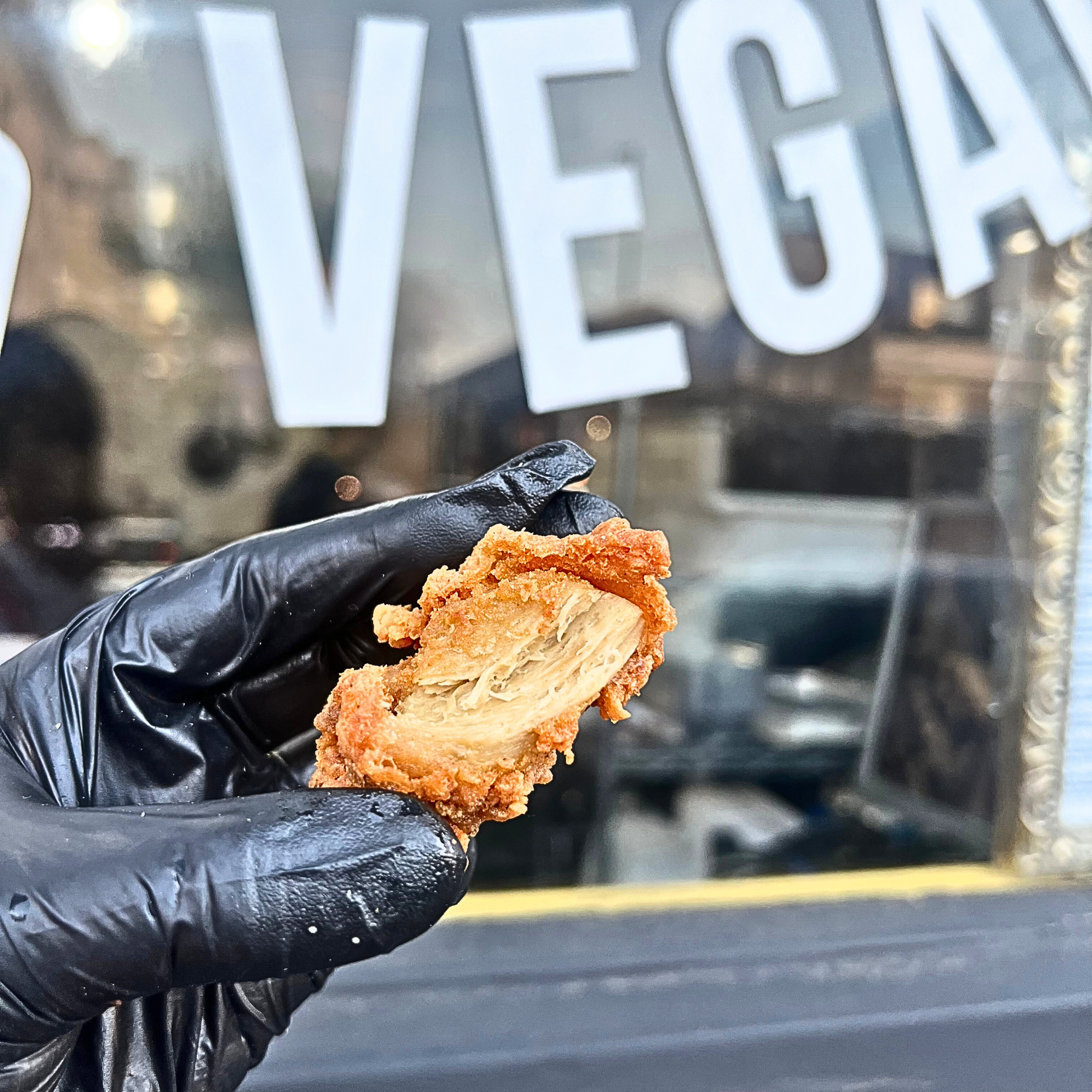 a hand with black gloves showing our vegan chicken realistic texture, it is stringy and juicy. It is has a crispy fried coat like KFC and juicy inside. There is also a big vegan sign behind, serving suggestion.